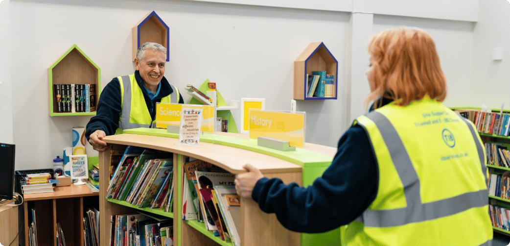 Colin and another TIB operative move a bookcase in a school library 