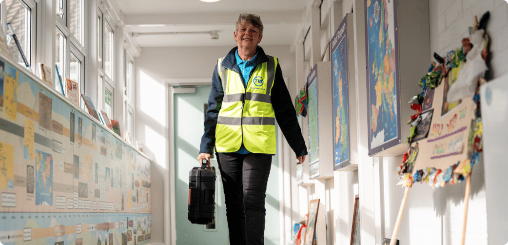 Jan walks through a school corridor while carrying a toolbox and wearing a TIB high vis