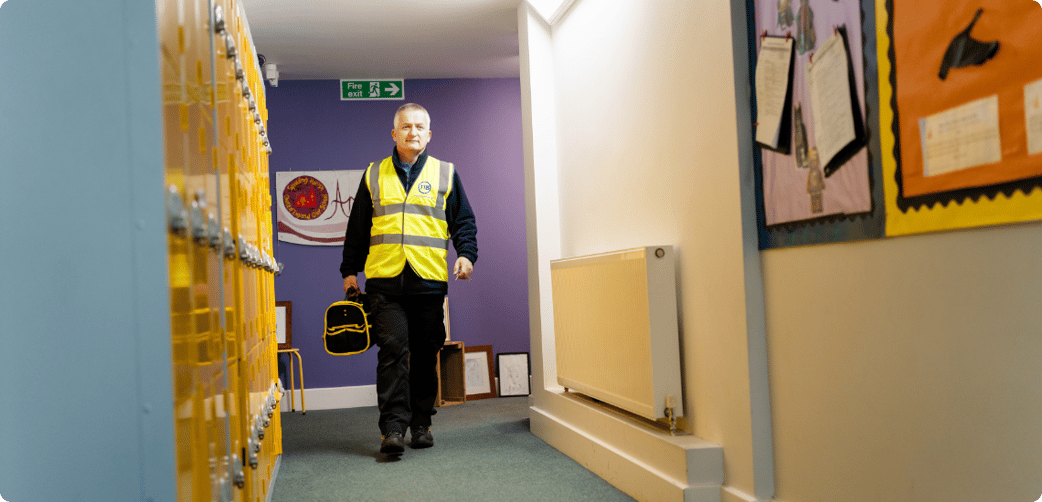 Leigh walks through a school corridor while carrying a toolbox and wearing a TIB high vis