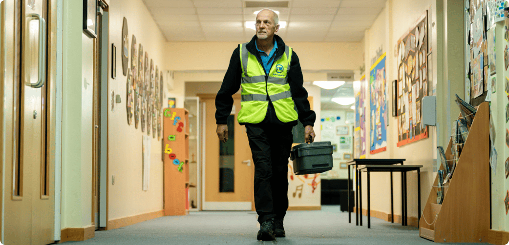 Ted walks through a school corridor while carrying a toolbox and wearing a TIB high vis