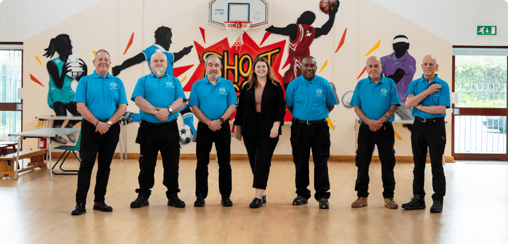 Ted stands with six other people in a school gym hall