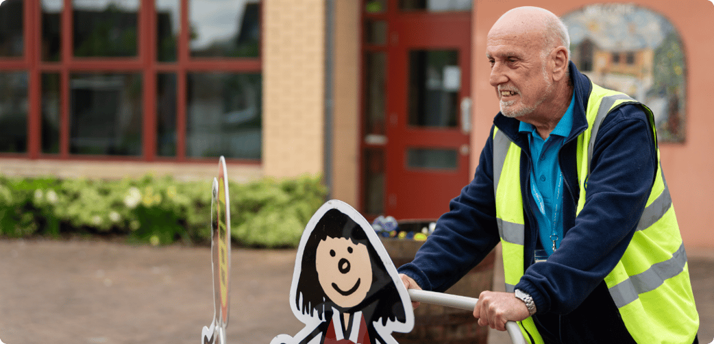 Tim pushes a sign past the entrance of a school while smiling and wearing a TIB high vis