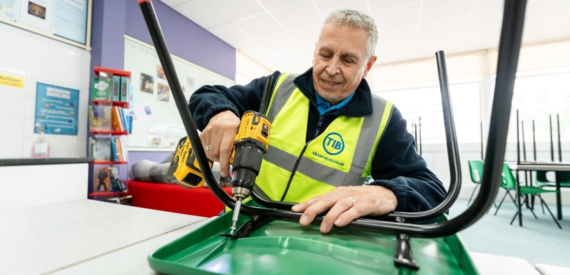 A TIB Services caretaker fixes a school chair while smiling