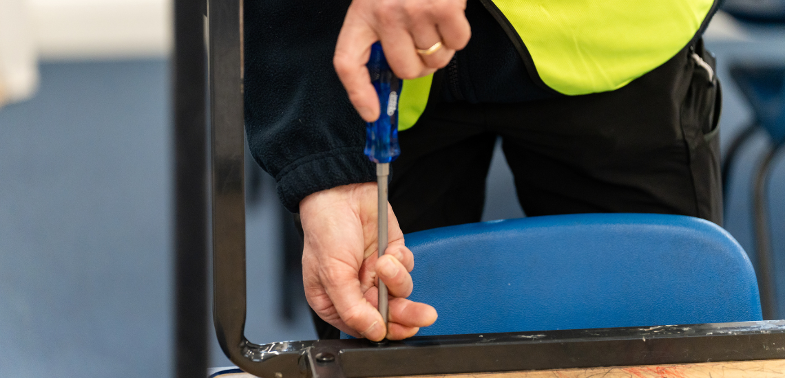 A TIB Services caretaker fixes a school desk with a screwdriver