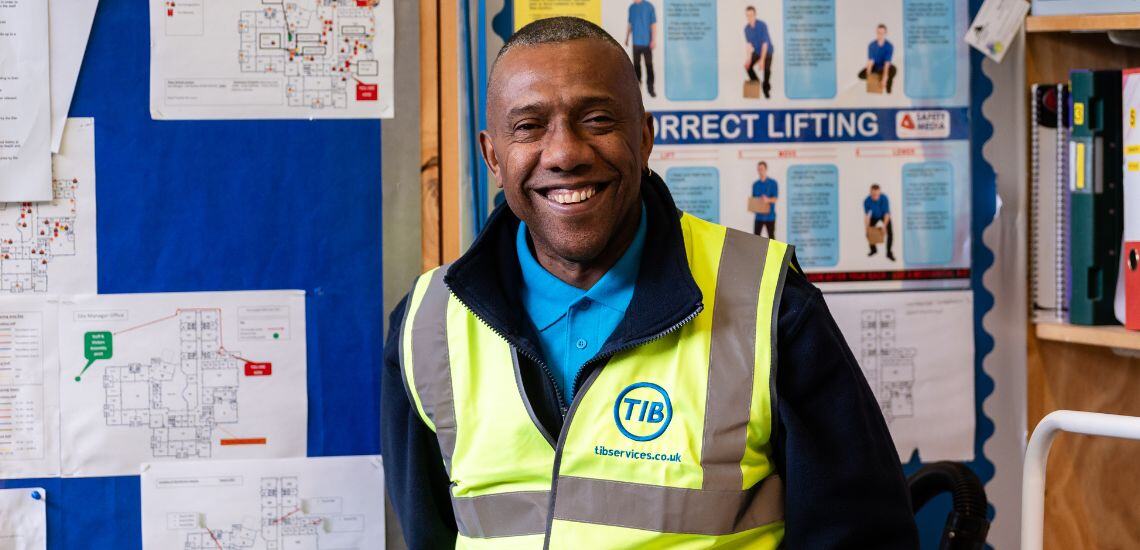 A TIB Services school caretaker stands in front of a notice board and smiles